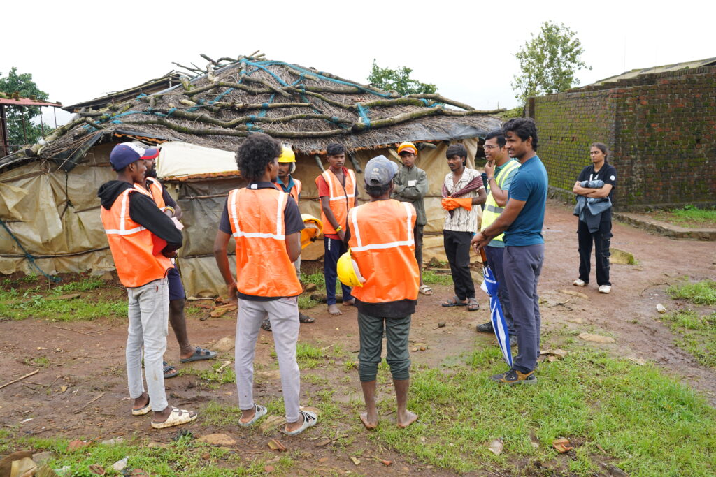 A group stands outdoors in front of makeshift shelters with tarpaulin roofs during a daytime meeting