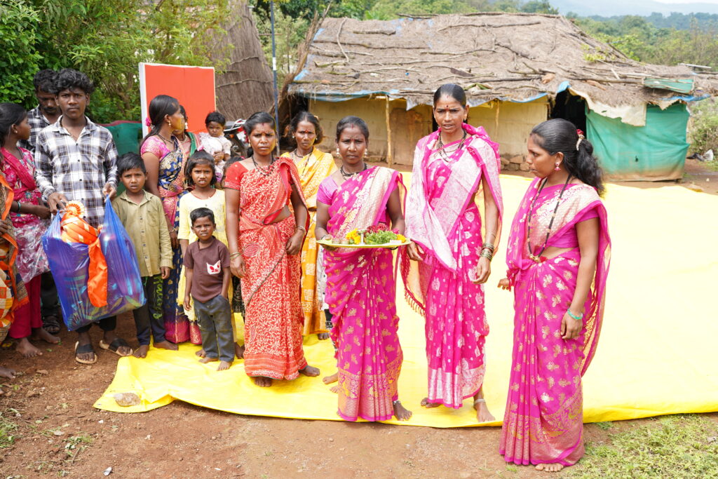 Residents in traditional dress standing on yellow mat in front of rural homes during daytime gathering