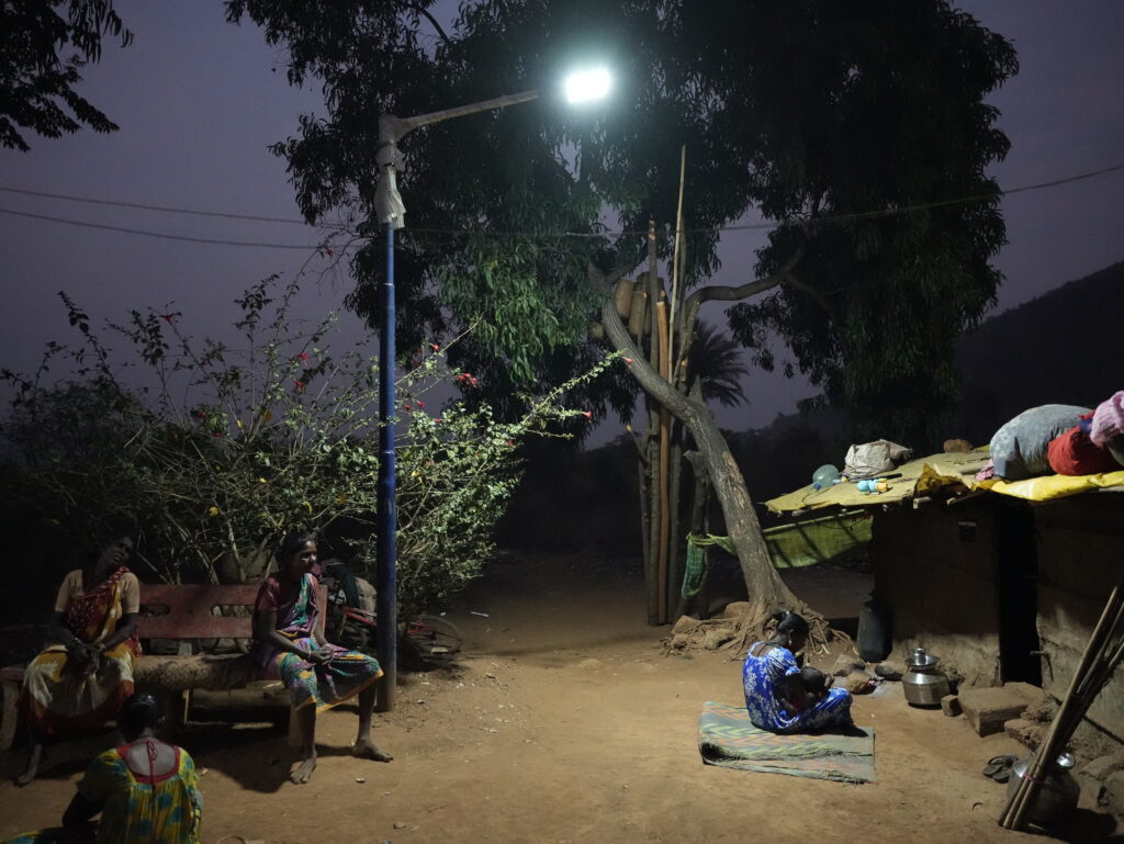 A solar streetlight illuminates an unpaved courtyard at dusk, with residents seated nearby
