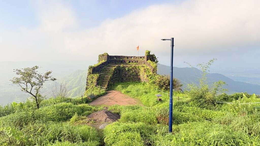Stone fort structure with flag on hilltop, blue solar street light pole in foreground, green hills and mountains in background