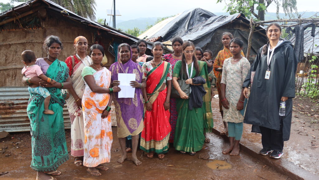 Women and field staff stand together outdoors in a hamlet, some holding papers