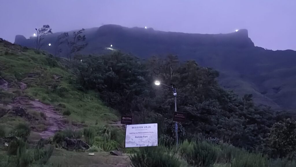 A solar streetlight pole lit at dusk on a hillside path with vegetation and mountain ridges visible in the background under a purple twilight sky