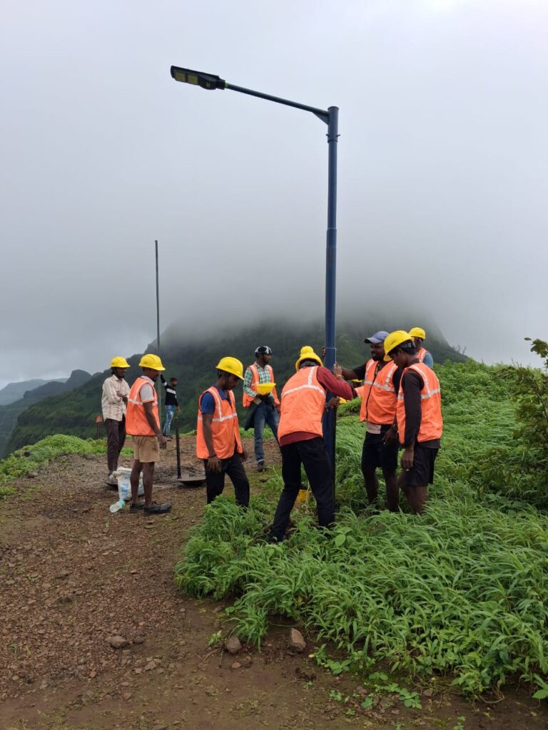 Installation crew in orange safety vests and yellow hard hats standing around a tall solar streetlight pole on a misty hilltop