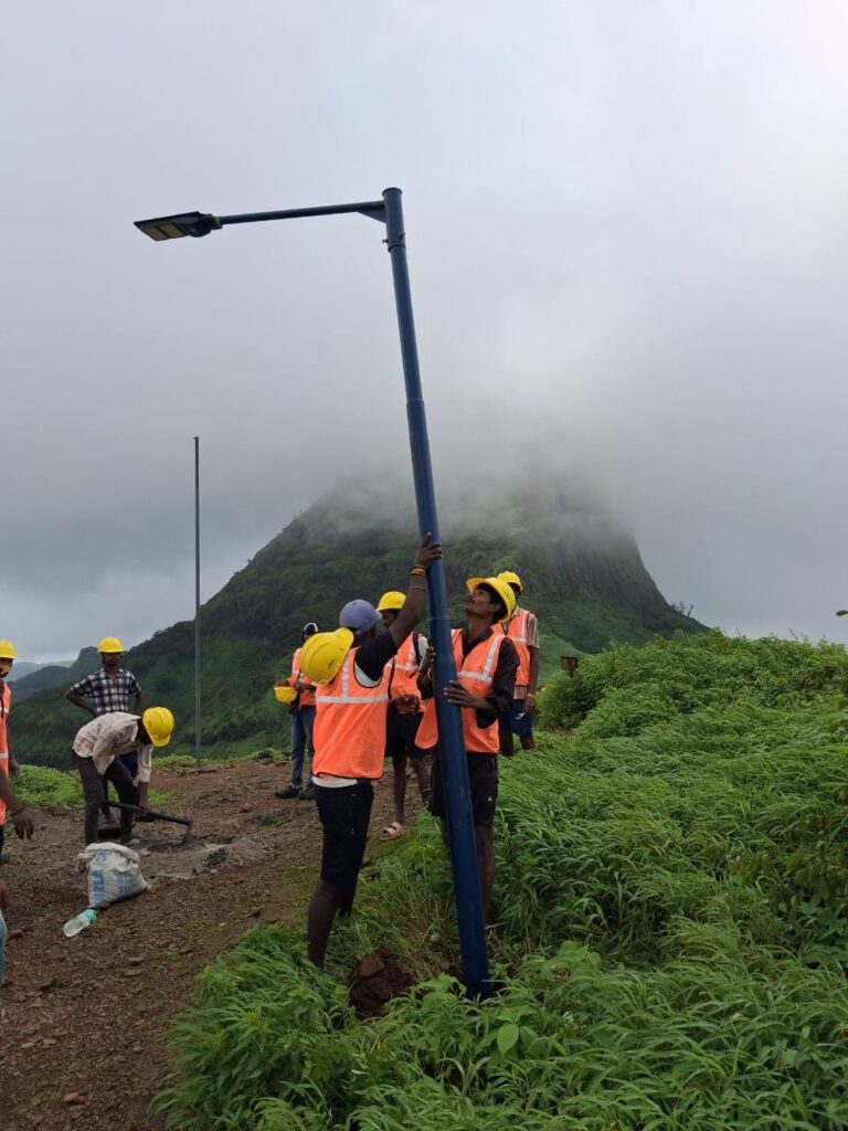 Workers in safety gear positioning a solar streetlight pole into the ground on a foggy hillside with mountain peaks shrouded in clouds behind them