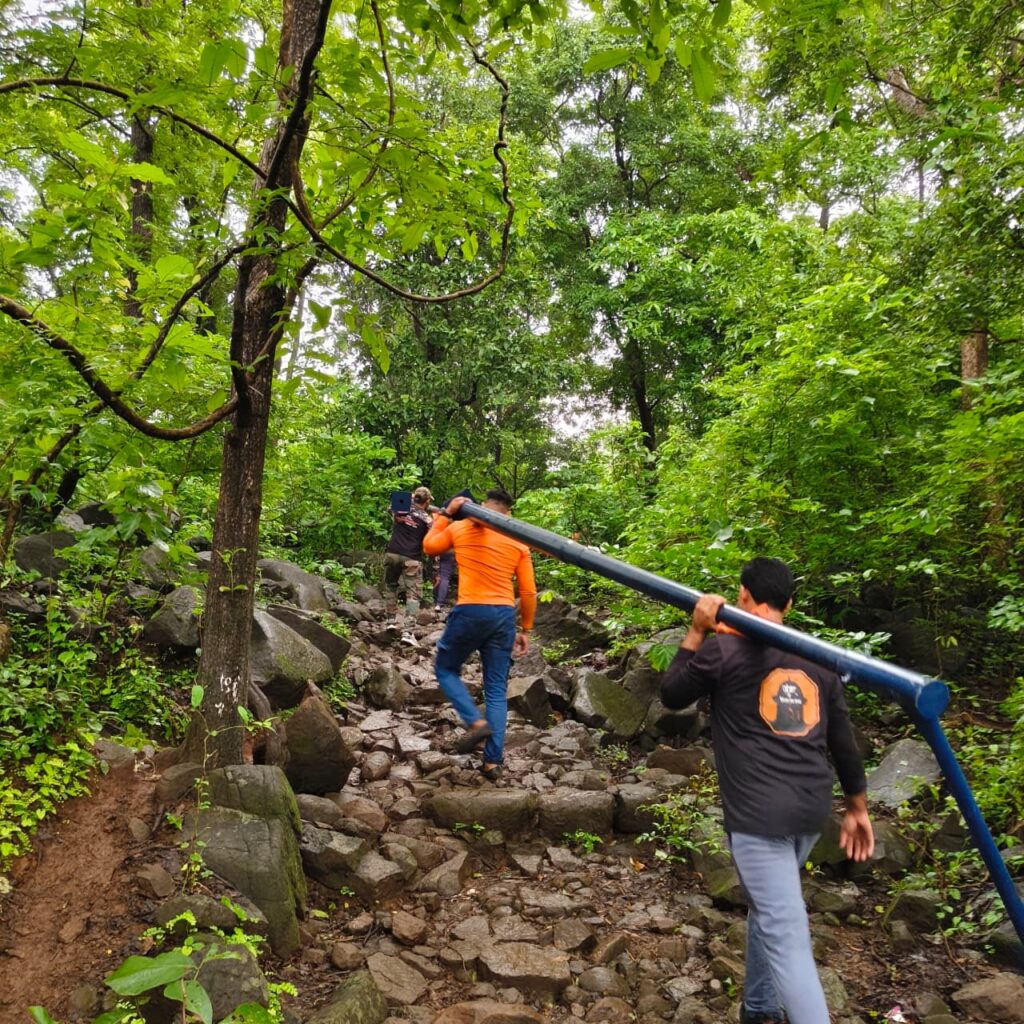 Installation team carrying a long pole through a forested rocky stairway with stone steps and dense green foliage overhead