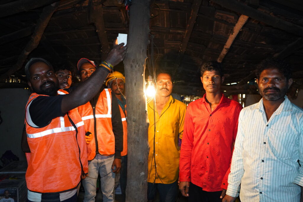 Residents stand inside a structure as a technician points to a lit bulb hanging from the ceiling