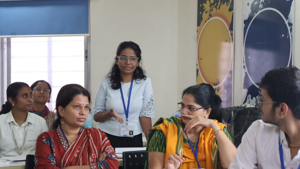 A woman standing and speaking to seated participants in a training room with windows and decorative wall art